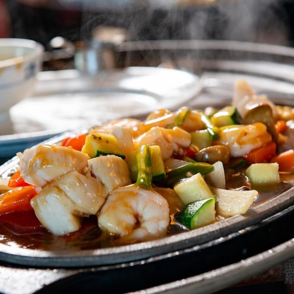 Sizzling Asian-fusion dish with shrimp, scallops, and vegetables on a hot plate, emitting steam.